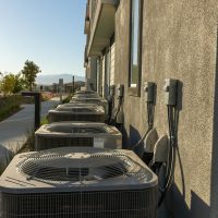 Line of air conditioning units on a city apartment exterior with a sidewalk view.