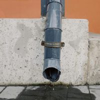 A close-up image of a metal drainage pipe on a concrete wall with visible water runoff.