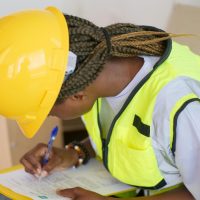 Design, Engineering & Project Management A female engineer in PPE writing on a clipboard at a construction site.