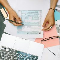 Design, Engineering & Project Management Hands writing on tax documents with laptop, glasses, and currency on desk.